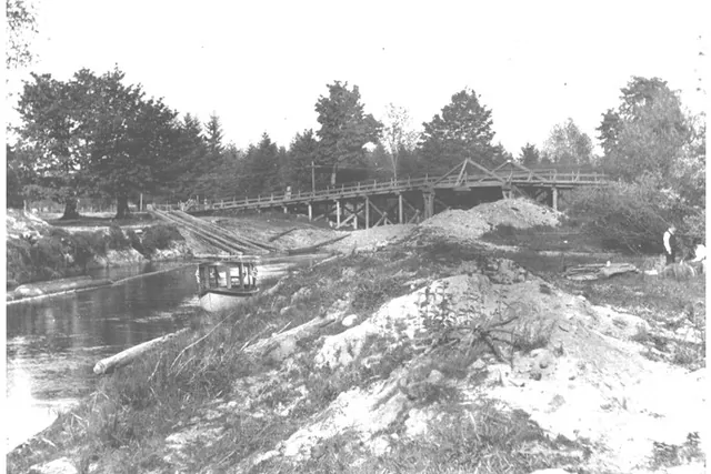 Black and white photograph of a narrow river with a long wooden trestle bridge behind it, a small boat on the water, and sandy banks.