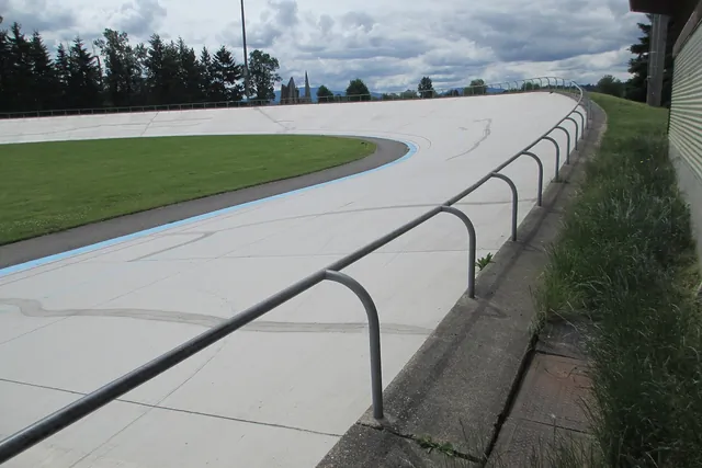 A pale concrete cycling track curves up into a steep bank around a green infield, with evergreen trees beyond.